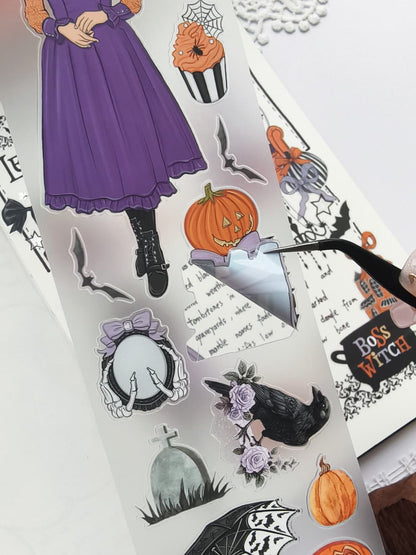 Close-up of tweezers peeling a die-cut Halloween PET sticker from the backing sheet, featuring spooky pumpkins, bats, a raven, skeleton mirror, gravestone, roses, and other gothic decorative elements.
