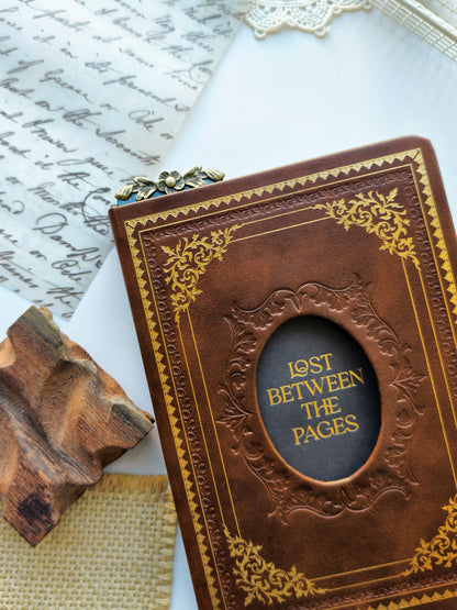 A brown faux-leather journal or notebook cover with ornate gold embossed detailing and a central oval cutout reading &