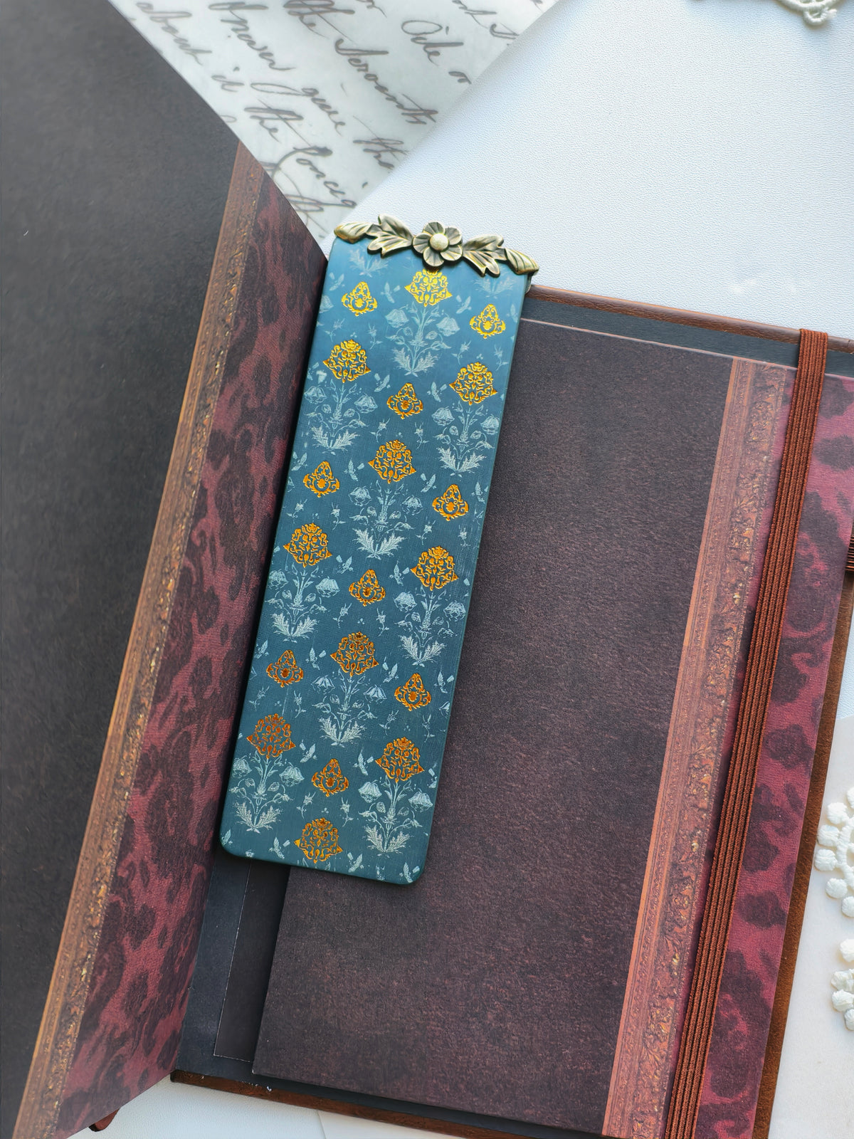 A decorative rectangular bookmark or card with a blue floral pattern and a small gold metal accent at the top, placed inside an open vintage-style book with dark maroon and gold detailing.