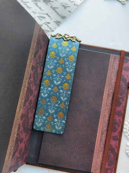 A decorative rectangular bookmark or card with a blue floral pattern and a small gold metal accent at the top, placed inside an open vintage-style book with dark maroon and gold detailing.