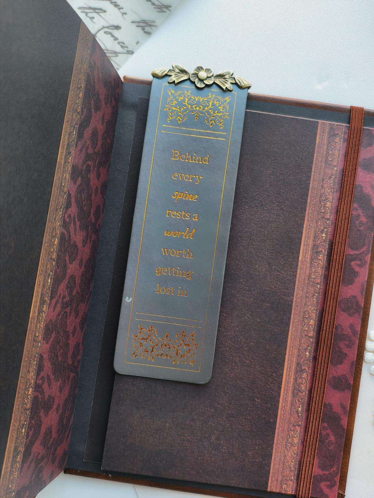 A dark gray rectangular bookmark with gold lettering and decorative borders, resting inside an open book with red and brown marbled pages.