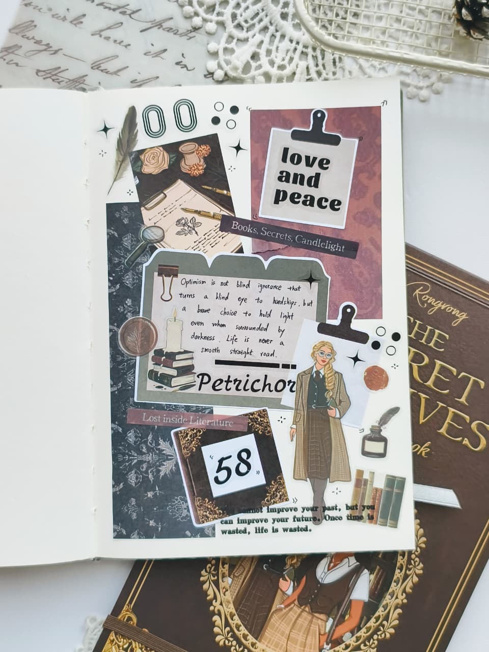 A decorative scrapbook or journaling page featuring vintage-style collage elements, including text boxes, clips, stars, a small illustrated figure, and book-themed decorations, photographed on top of books with a lace tray visible in the background.