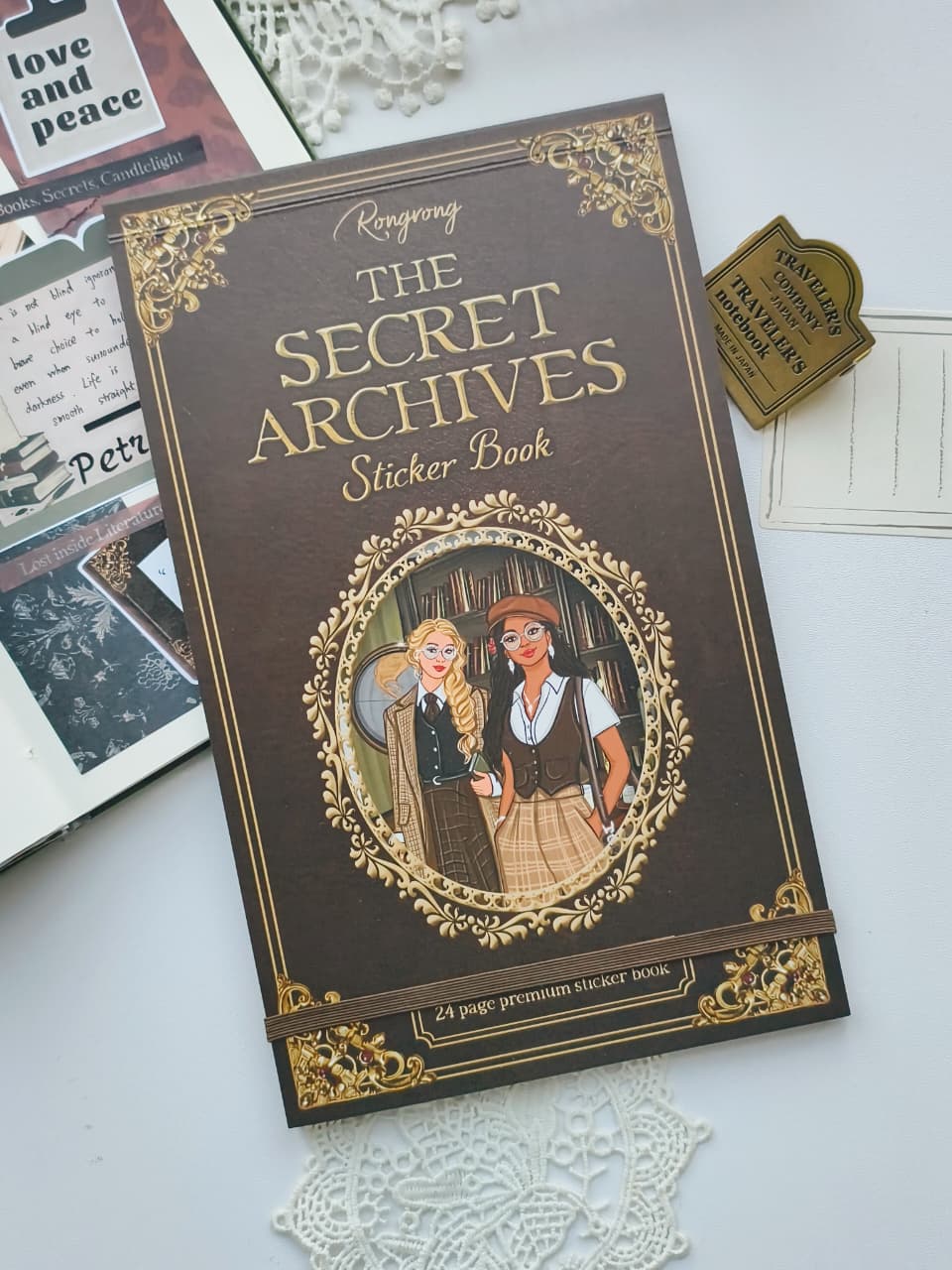 The Secret Archives Sticker Book, features a dark brown cover with ornate gold borders and an illustration of a couple in an oval frame. The book is styled on a white surface with vintage paper cards and lace doilies.