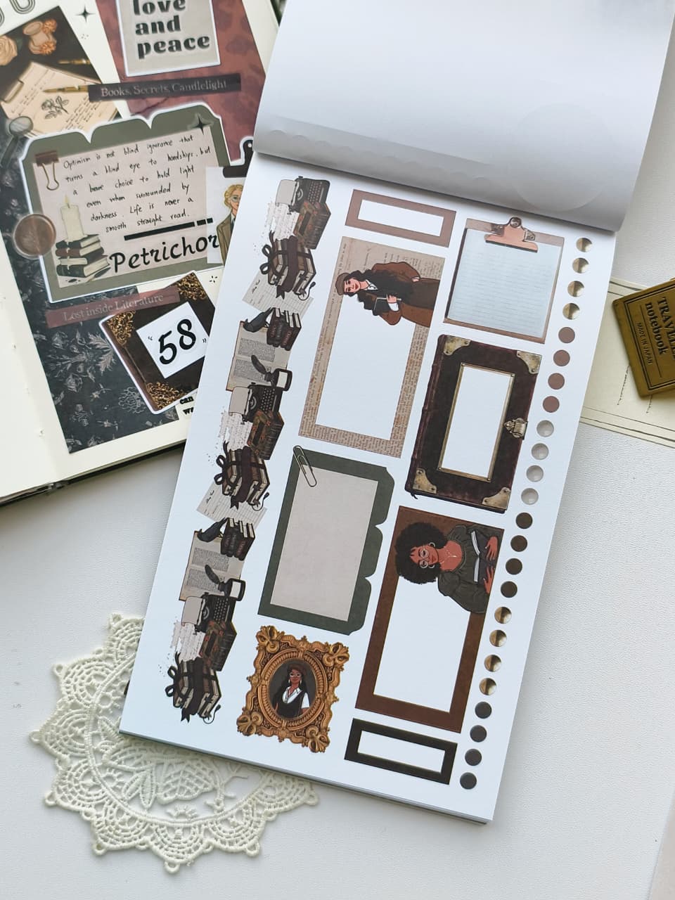 A sheet of vintage-themed planner stickers displayed on a white surface next to an open scrapbook journal. The stickers feature antique frames, sepia-toned illustrations, and decorative text elements.