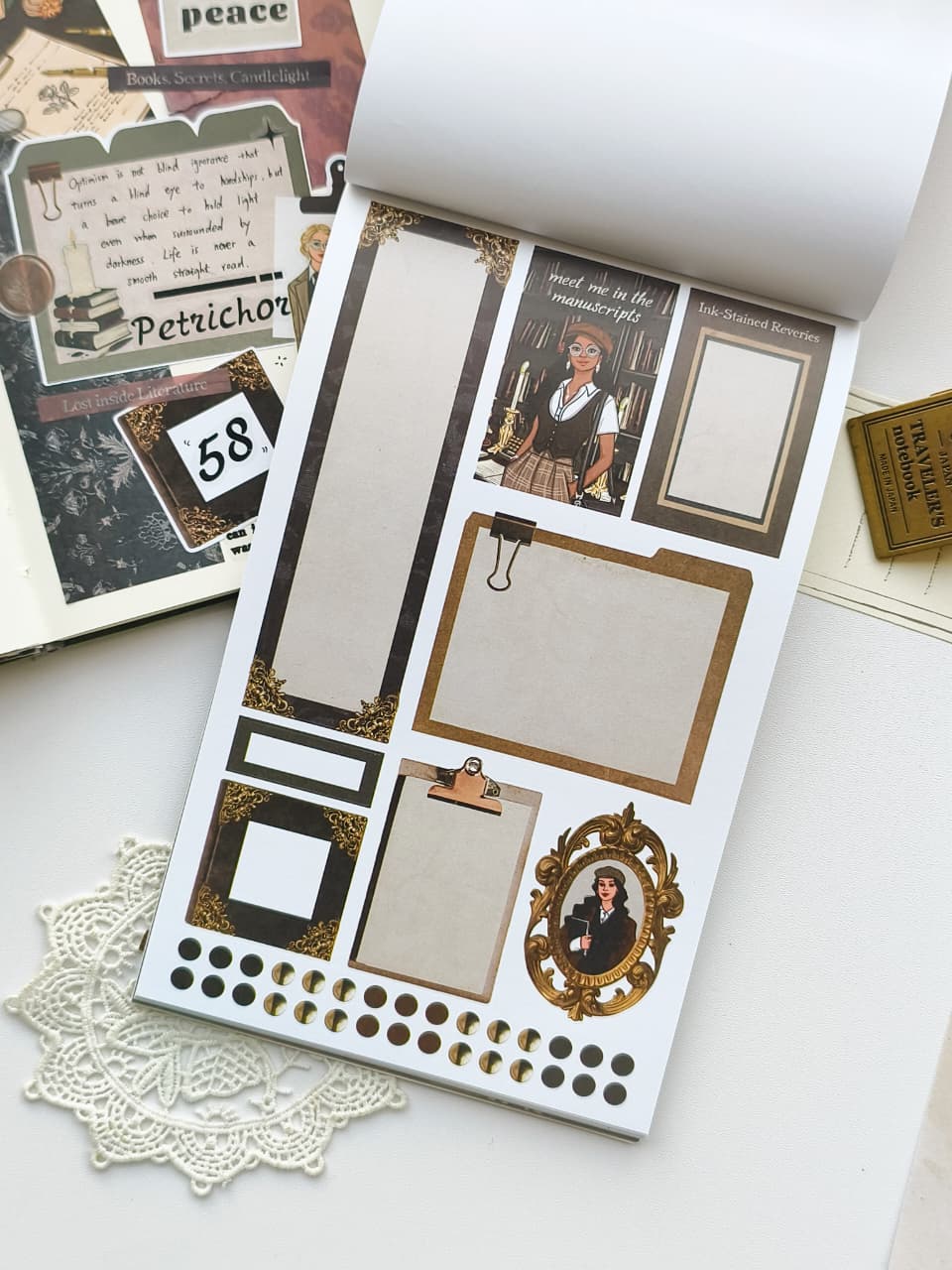 A sheet of vintage-style scrapbook stickers displayed on a white surface, surrounded by coordinating paper ephemera, a lace doily, and a small gold-toned box. The stickers include ornate frames, text boxes, clipboards, a portrait frame, and decorative dots.