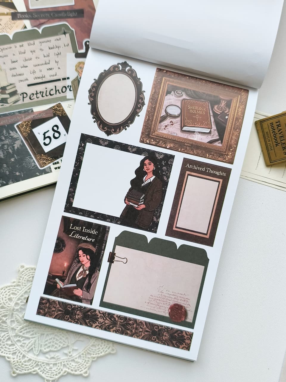 A styled flat-lay of vintage-themed scrapbook or journaling stickers on a white sheet, surrounded by matching ephemera, a lace doily, and a book page.