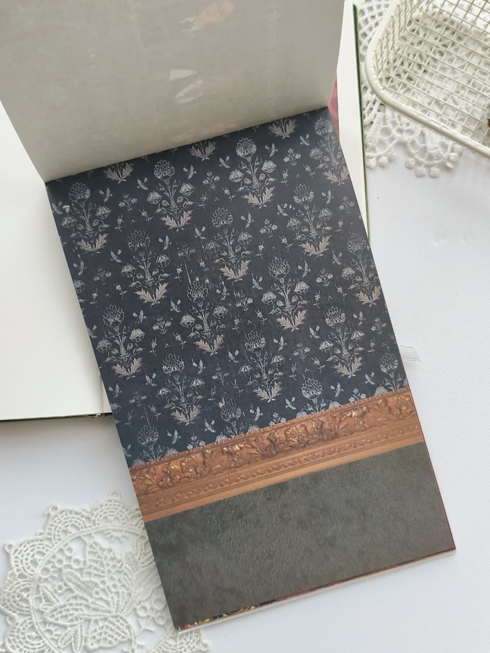 A folded book or notebook with a dark navy floral patterned cover and a decorative gold border, photographed on a white surface with lace and a woven basket nearby.