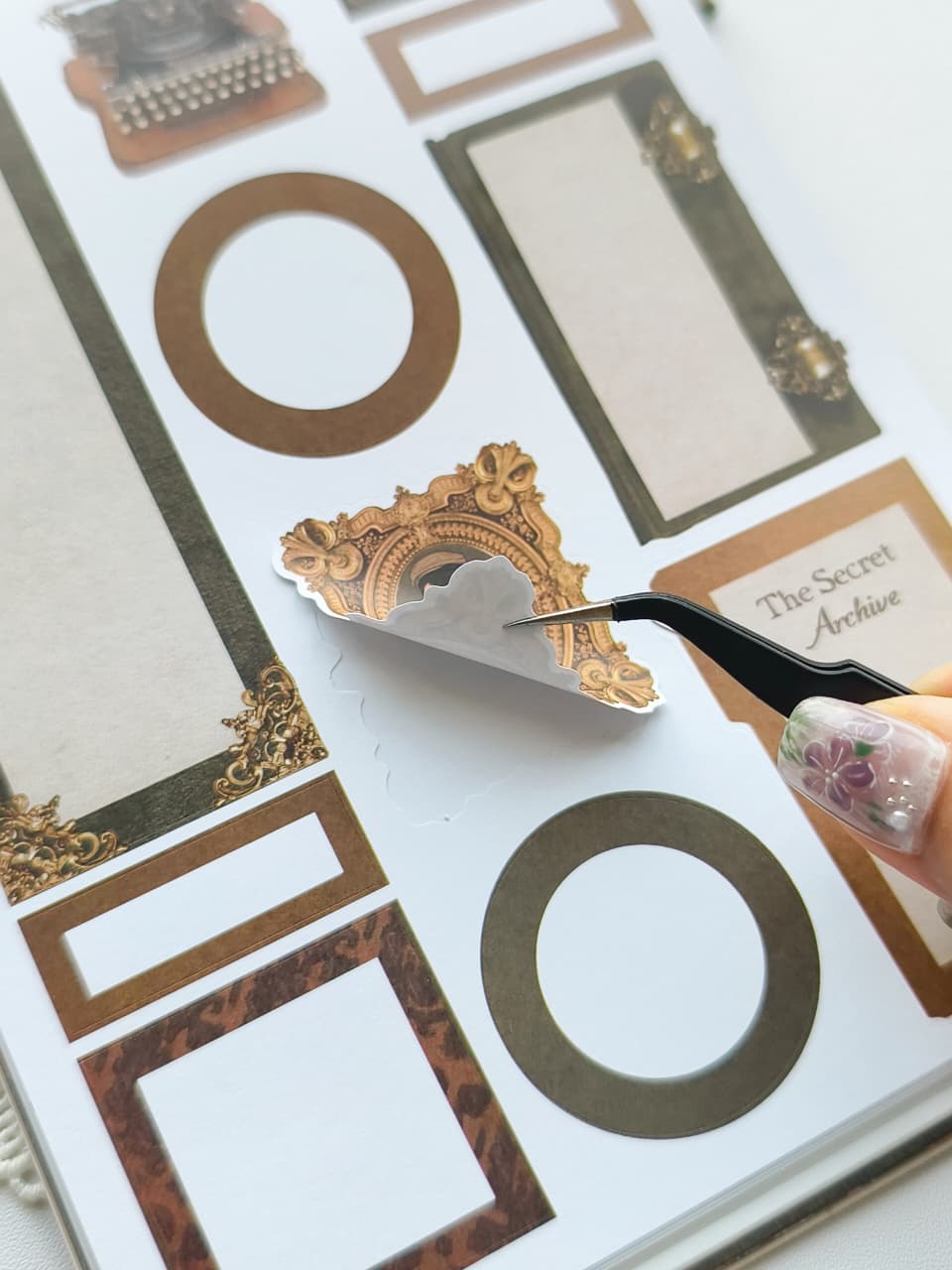 A close-up of a hand using black tweezers to peel a decorative gold and white ornate sticker from a sheet of planner stickers. The sheet contains various gold, brown, and beige vintage-style frames, circles, and labels, including one that reads 'The Secret Archive'.