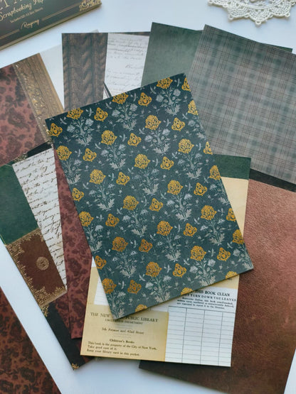 A styled flat-lay of vintage-themed scrapbooking papers and cardstock sheets arranged on a white surface. The central sheet features a dark teal floral pattern with yellow roses and white leaves, surrounded by other patterned papers in brown, green, beige, and plaid designs.