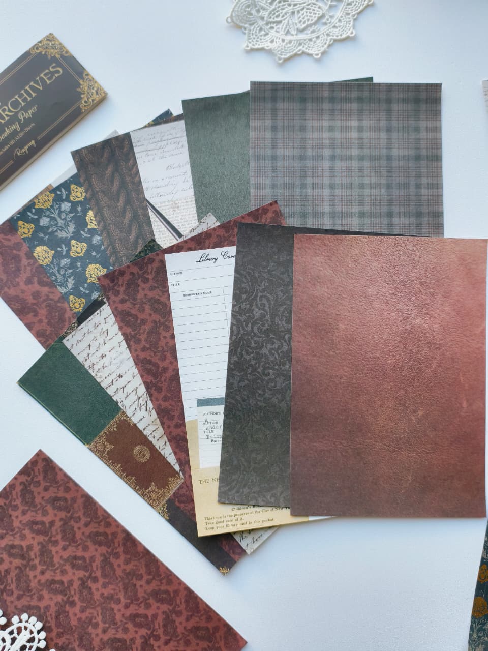 A flat-lay display of vintage-style scrapbooking papers and envelopes in various patterns, including plaid, floral, damask, and textured leather-like finishes in muted red, green, brown, and gray tones.