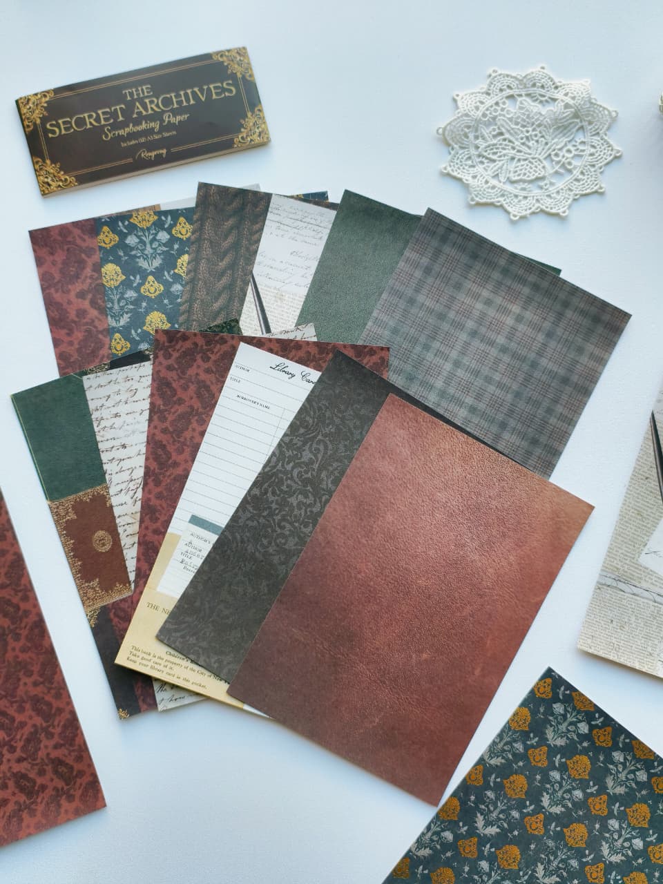 A flat-lay display of vintage-themed paper sheets and envelopes from The Secret Archives, featuring various patterns like plaid, damask, floral, and lined script in muted earthy tones, arranged on a light blue background alongside a white lace doily.