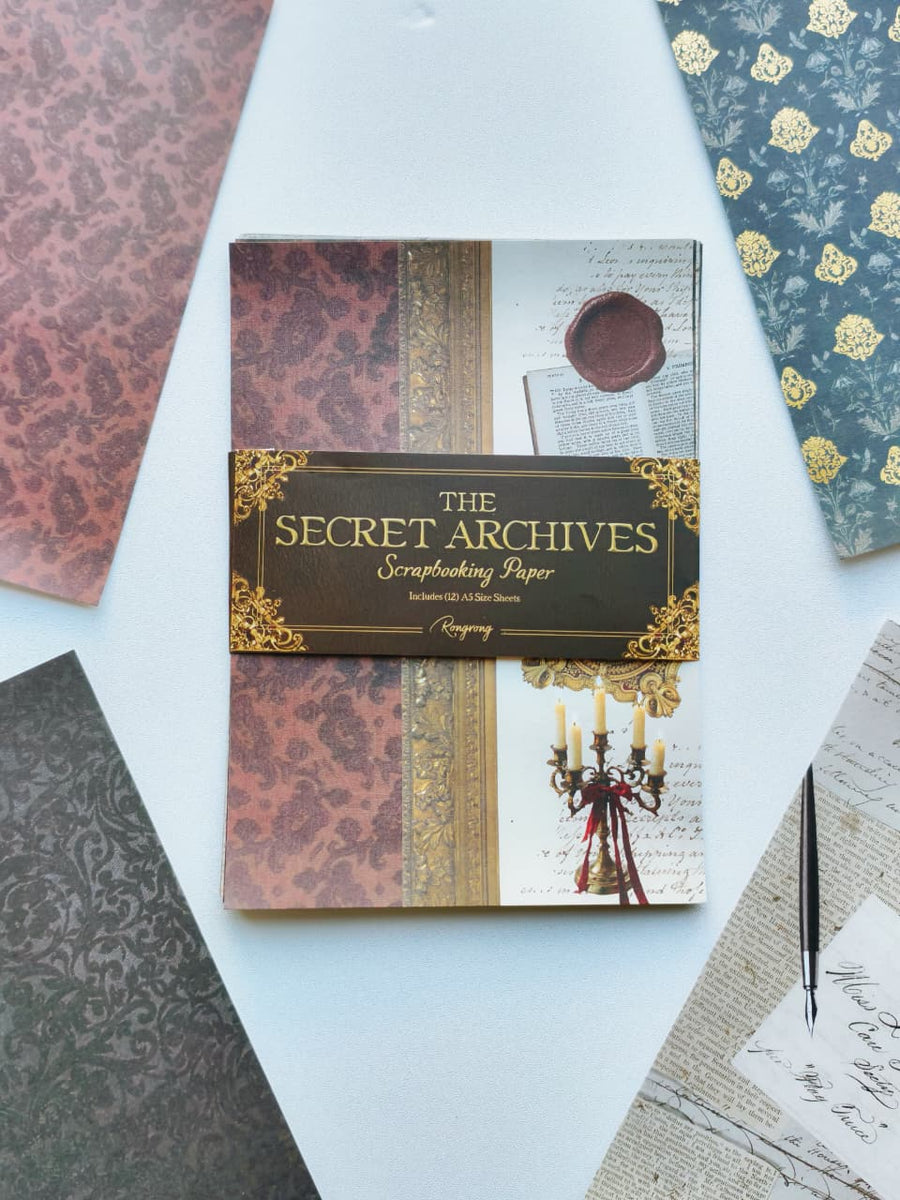 A flat-lay presentation of 'The Secret Archives' scrapbooking paper pack by Paperhouse, featuring a central pack of patterned papers wrapped in a decorative black and gold paper band, surrounded by other patterned papers on a white background.