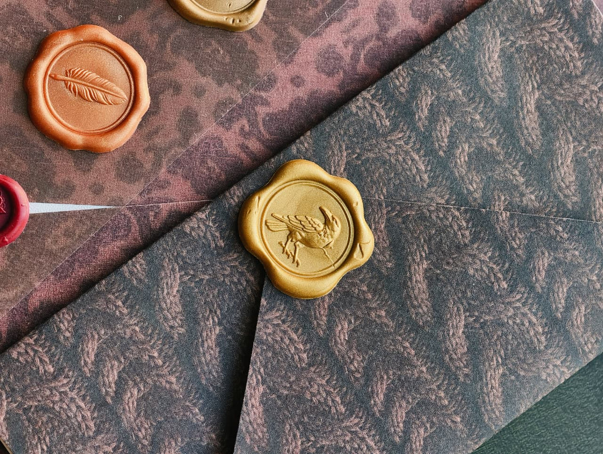 A close-up styled flat lay of patterned envelopes and wax seals. A dark charcoal-gray envelope with a leaf pattern is layered over a dusty rose envelope with a leopard print, both featuring embossed leaf motifs.