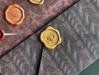 A close-up styled flat lay of patterned envelopes and wax seals. A dark charcoal-gray envelope with a leaf pattern is layered over a dusty rose envelope with a leopard print, both featuring embossed leaf motifs.