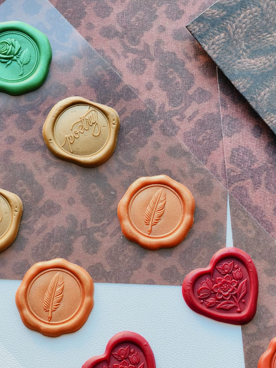 A styled flat-lay of wax seals in multiple colors, including green, gold, orange, and red, arranged on patterned brown paper and a white surface.
