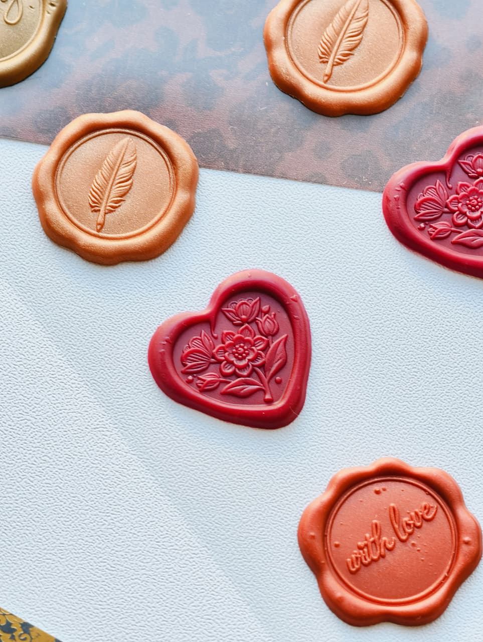 Decorative wax seals arranged on a light textured surface, including round copper-toned seals with feather and script motifs, a red heart-shaped floral seal, and another red seal with a flower border.
