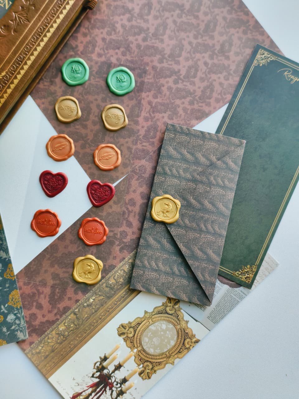 A styled flat-lay of vintage wax seals in green, gold, orange, and red, arranged on patterned brown paper alongside a gray patterned envelope with a gold seal, decorative cards, and a gold frame.