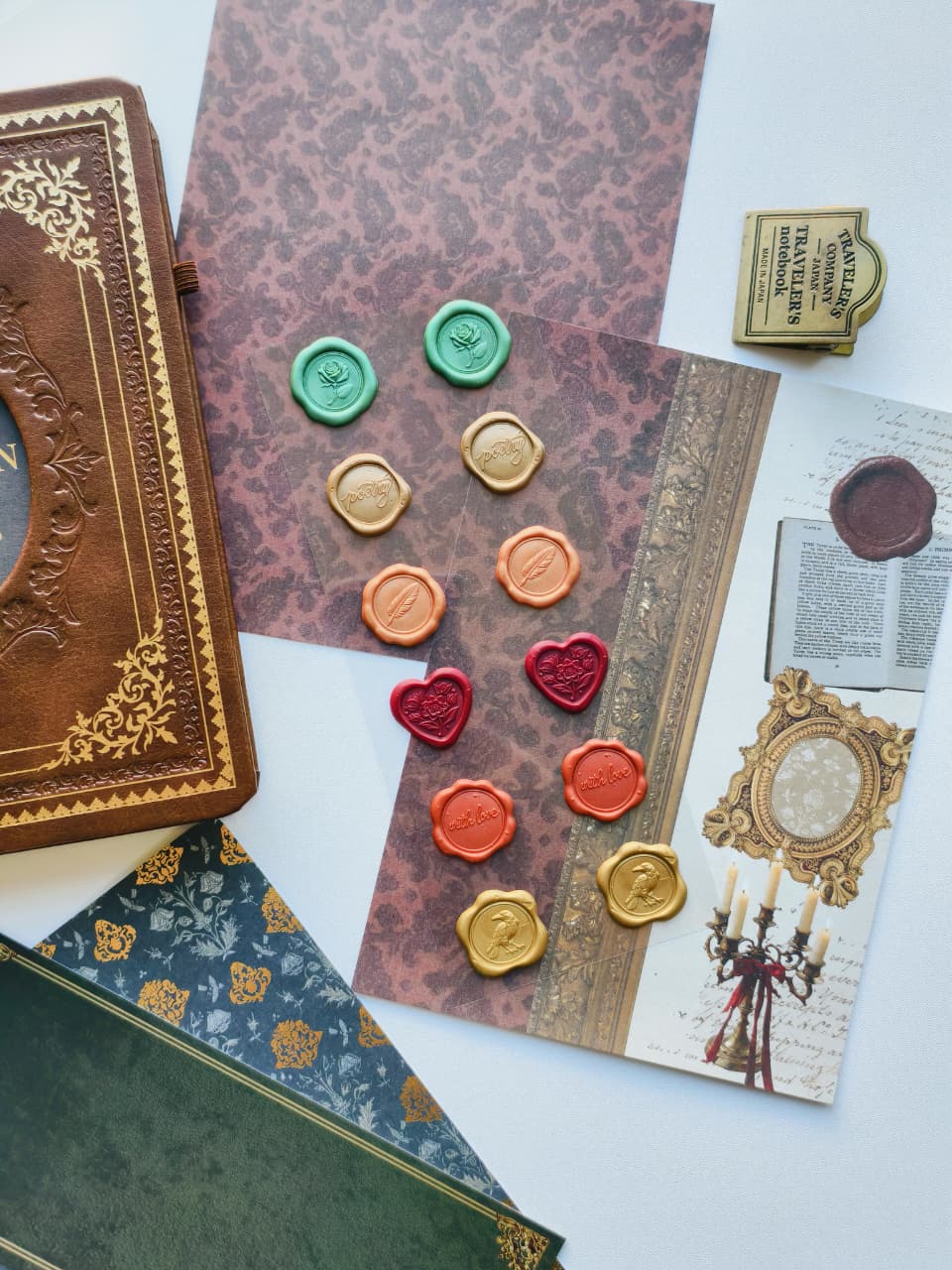 A styled flat-lay of vintage-themed stationery and decor items on a white surface, including patterned envelopes, an embossed brown journal, wax seals in green, gold, orange, and red, a small brass stamp, and printed cards with antique illustrations.