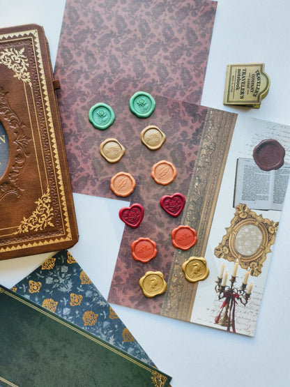 A styled flat-lay of vintage-themed stationery and decor items on a white surface, including patterned envelopes, an embossed brown journal, wax seals in green, gold, orange, and red, a small brass stamp, and printed cards with antique illustrations.