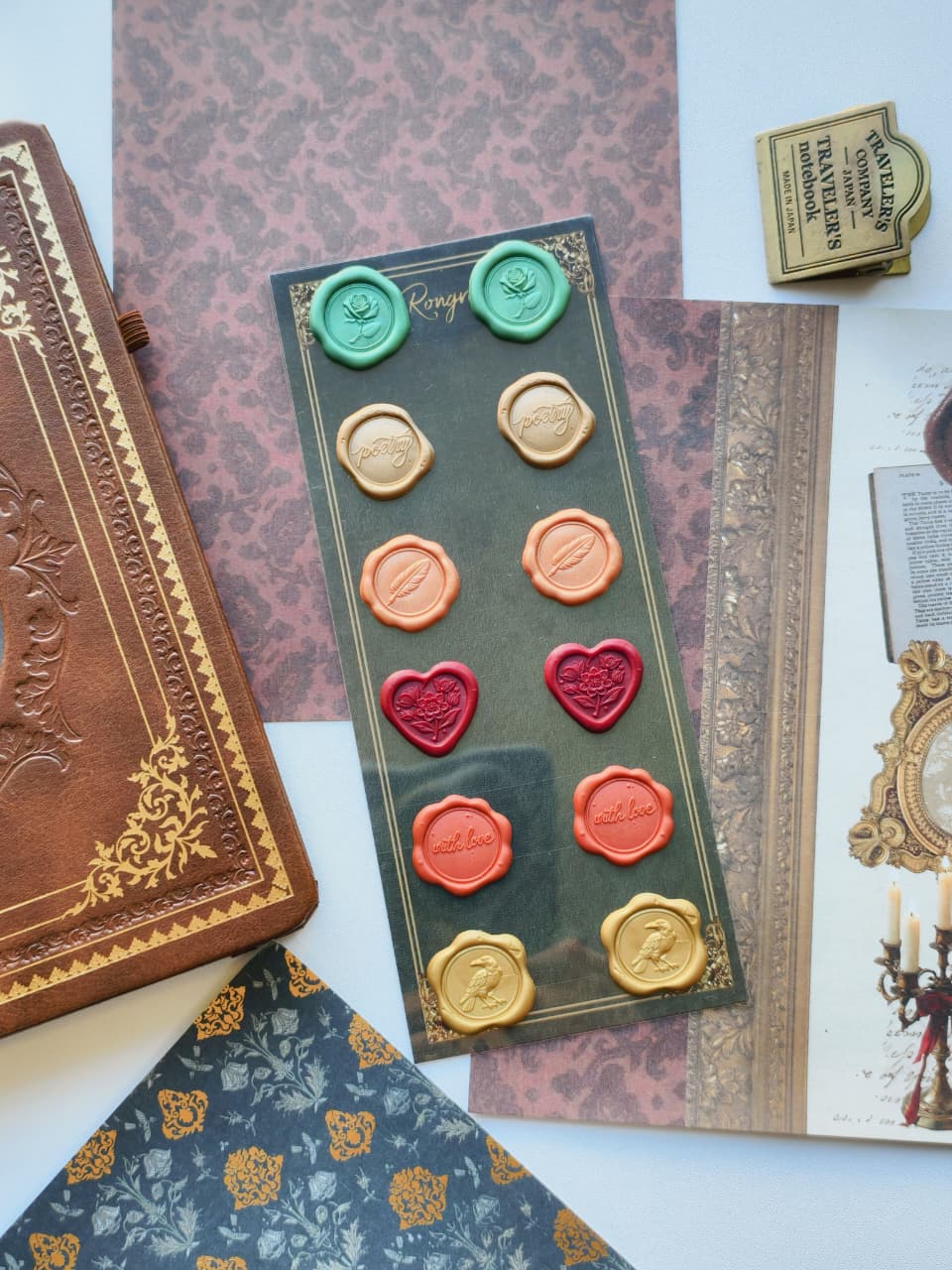 A wax seal stamp set displayed on a dark green card, surrounded by vintage-style props including an embossed brown journal, patterned papers, and an open book. The set includes multiple wax seals in gold, green, peach, red, and orange, along with two matching stamps.