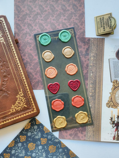 A wax seal stamp set displayed on a dark green card, surrounded by vintage-style props including an embossed brown journal, patterned papers, and an open book. The set includes multiple wax seals in gold, green, peach, red, and orange, along with two matching stamps.